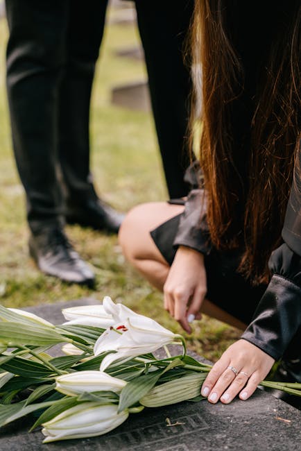 A woman with long brown hair, dressed in dark clothing, kneeling on the grass and arranging a floral bouquet of white lilies with green leaves. The lilies are fresh, with smooth, white petals and prom