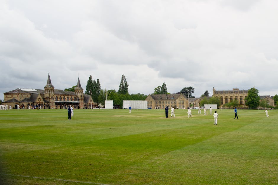 A wide view of a cricket field near Lords Cricket Ground, with players dressed in white and dark uniforms positioned on the lush green pitch. The background features historic buildings, including a ca
