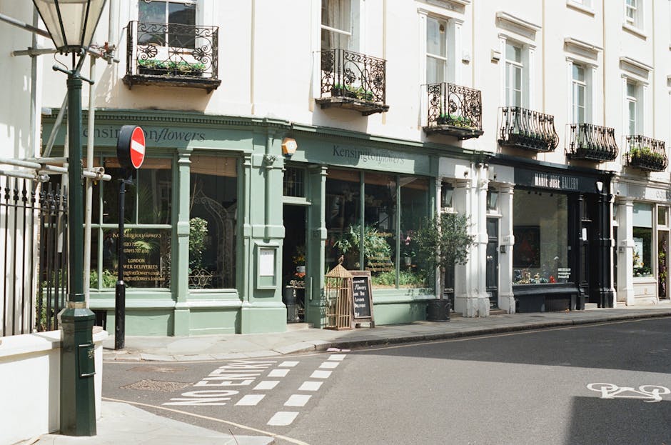A street view outside a flower shop with a green storefront, large display windows showcasing various floral arrangements, and a blackboard sign on the sidewalk. The shop, part of St Johns Wood floris