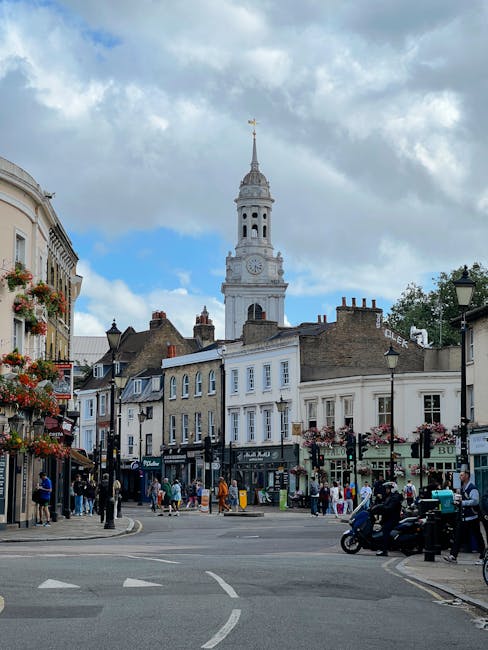 A busy street scene in St Johns Wood with rows of charming, white-painted and beige brick buildings, some adorned with hanging flower baskets. In the background, the prominent white clock tower with a